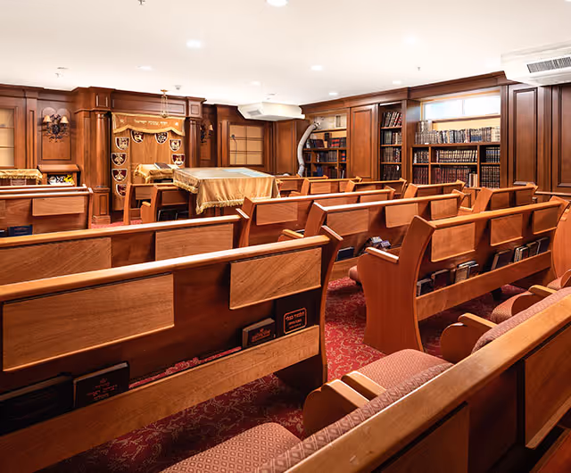 Wood-paneled chapel or synagogue interior with rows of wooden pews, bookcases, and an ark at the front.