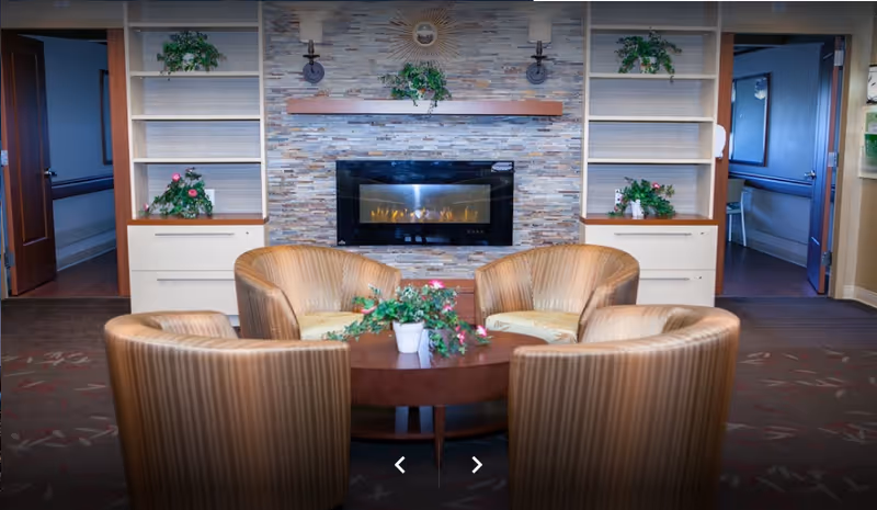 A cozy sitting area with four striped armchairs arranged around a round wooden table with a potted plant. Behind the seating area is a modern electric fireplace set in a stone wall with built-in shelves on either side, decorated with green plants. Two open doorways are visible on each side of the fireplace.