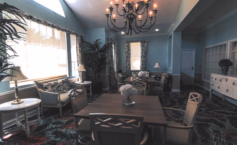 A softly lit dining room with light blue walls, floral curtains, and a large window letting in natural light. The room features several wooden tables and chairs, a chandelier hanging from the ceiling, potted plants, table lamps, and a sideboard with decorative items.