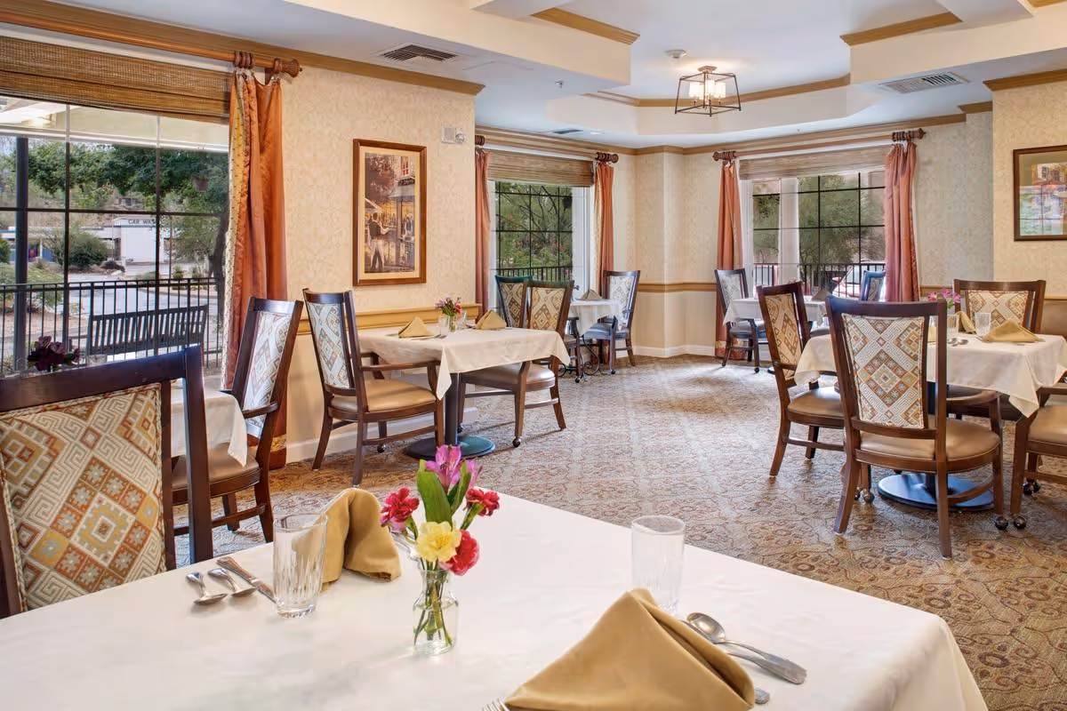 Bright dining room with multiple tables and upholstered chairs set with white tablecloths, napkins, glassware and small floral centerpieces, with large windows and curtains.