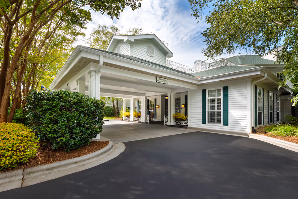 Exterior view of Arbor Terrace Asheville showing the covered entrance with white columns, green shutters on windows, and surrounding greenery including bushes and trees under a partly cloudy sky.