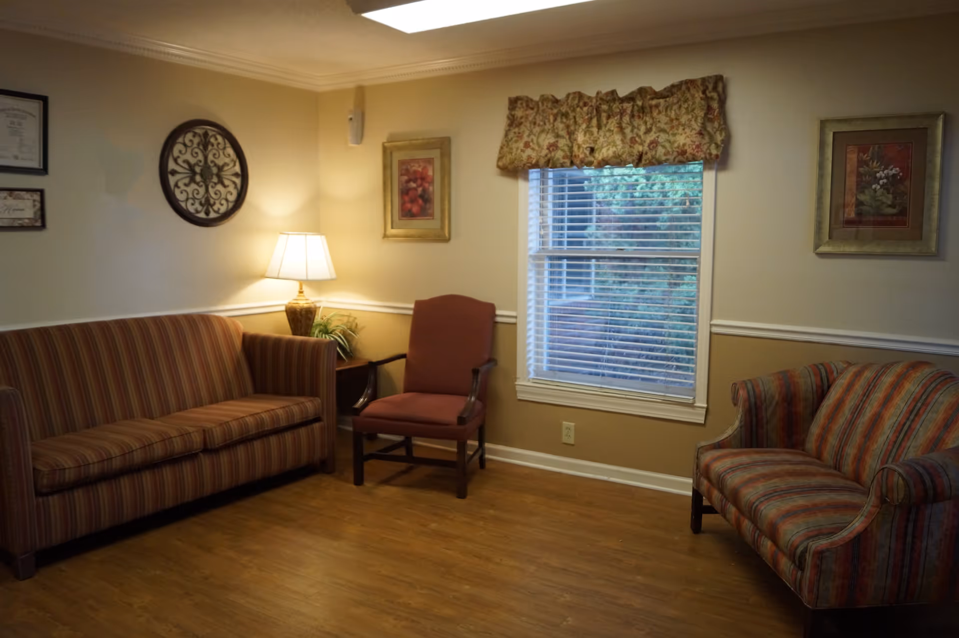 A cozy living room with two striped sofas and a red armchair arranged around a wooden floor. There is a side table with a lamp and a small plant, and the walls are decorated with framed artwork and a decorative wall piece. A window with floral valance and blinds lets in natural light.