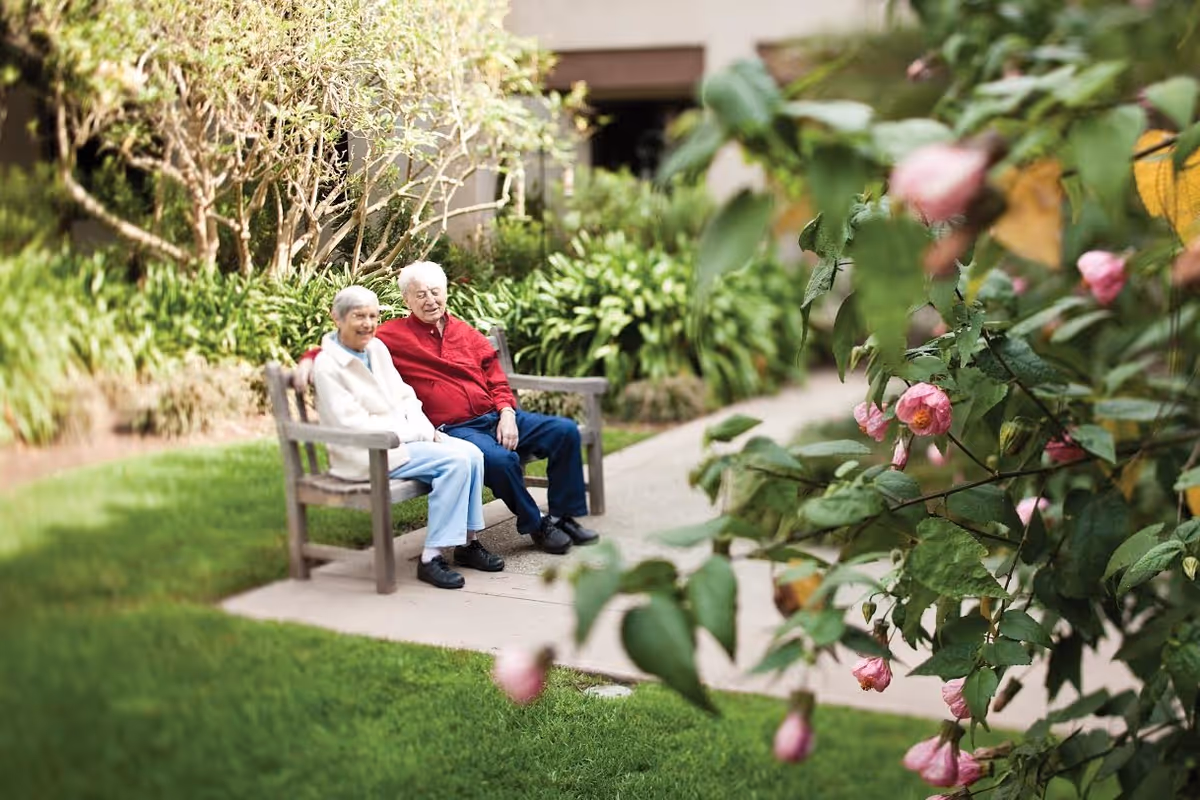 Two elderly people sitting on a wooden bench in a garden area with green grass, plants, and pink flowers in the foreground.