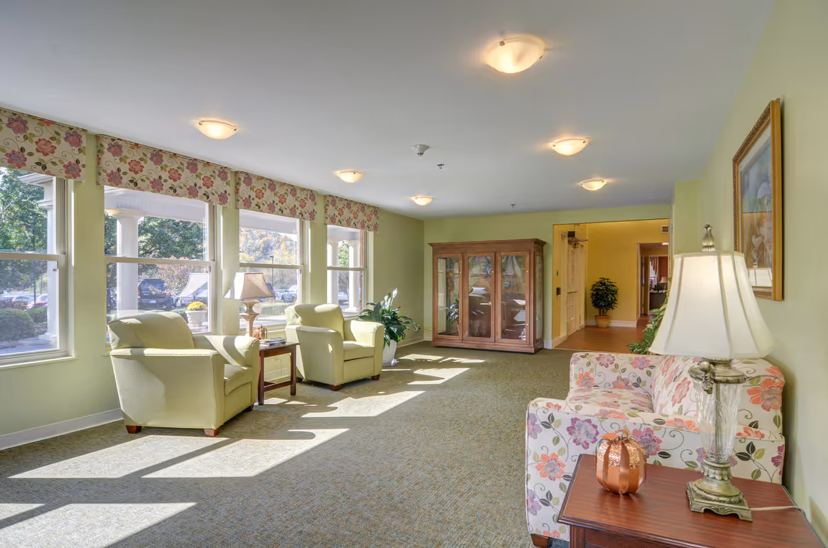 Sunlit common lounge with armchairs, floral sofa, side tables, lamps and large windows.