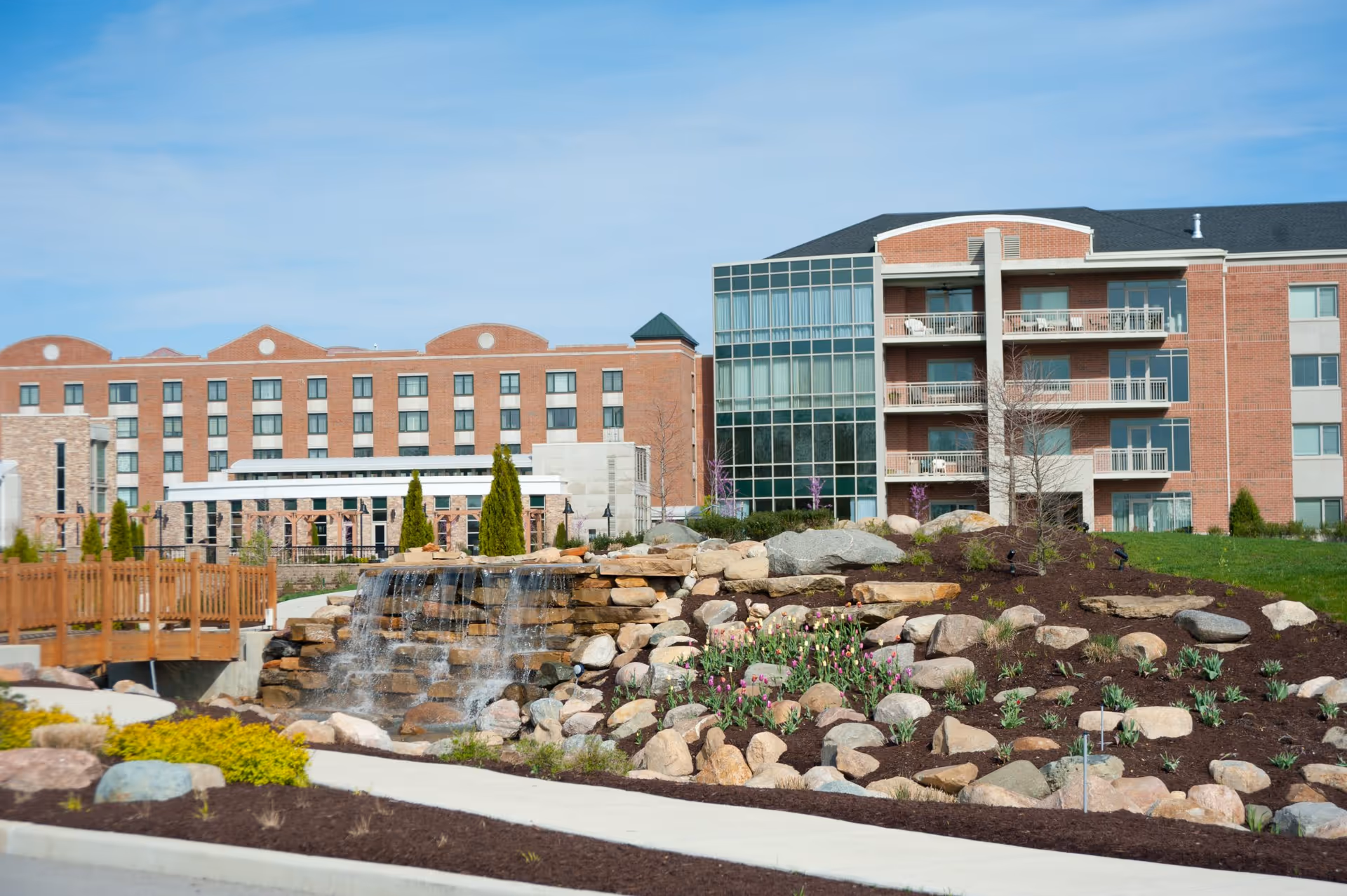 Outdoor view of a senior living facility named Marquette featuring a landscaped garden with rocks, flowers, and a small waterfall. The building is a multi-story brick structure with balconies and large windows under a clear blue sky.