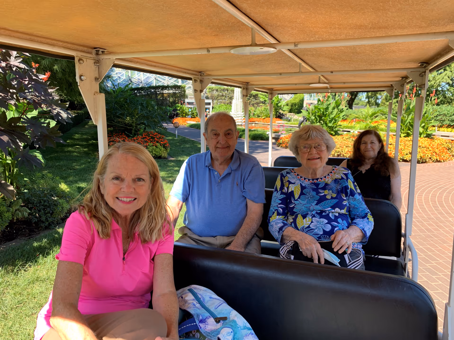 Four elderly people sitting on a covered outdoor tram or shuttle, surrounded by a garden with green plants and colorful flowers on a sunny day.