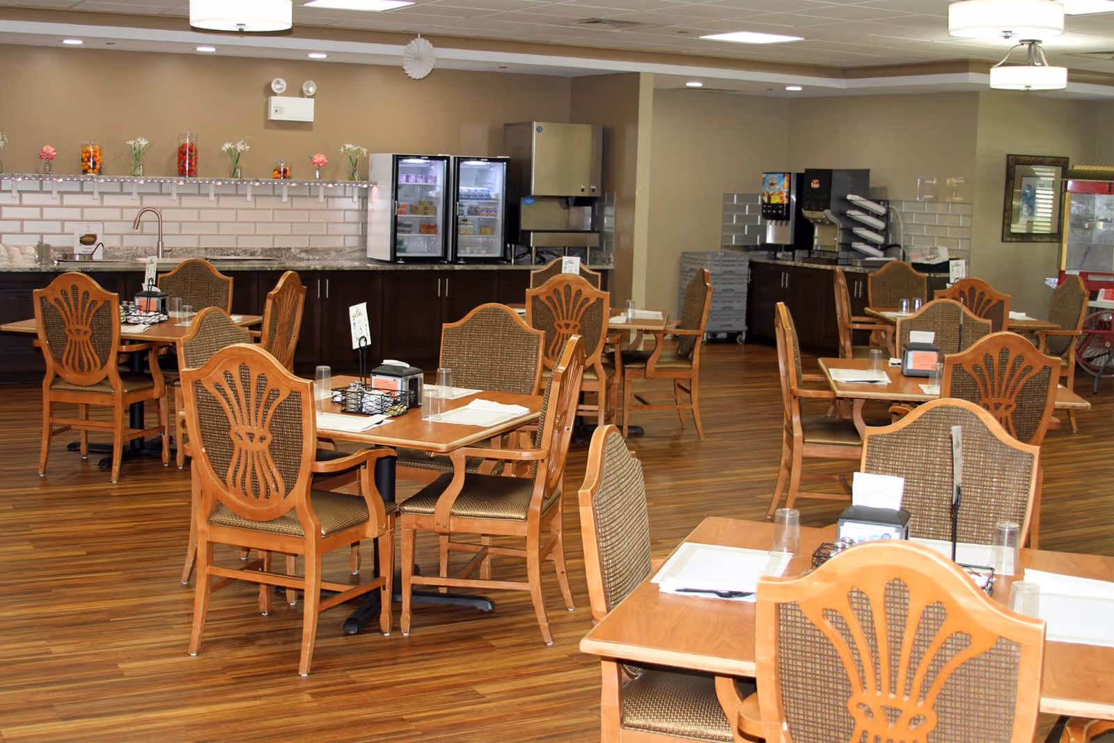 Spacious senior dining room with wooden tables and chairs and a serving counter with beverage machines in the background.