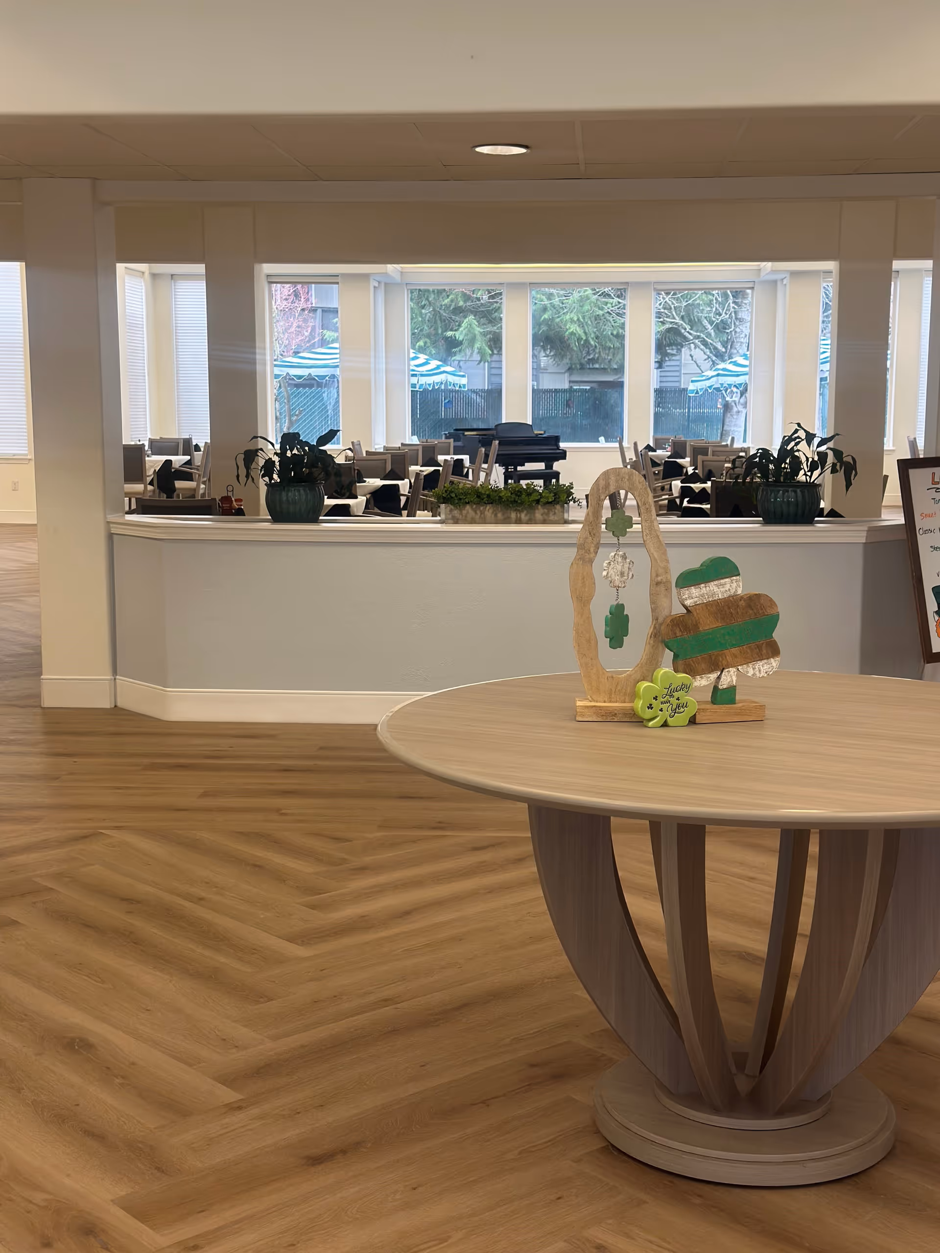 Interior view of a senior living facility with a round wooden table in the foreground decorated with St. Patrick's Day themed ornaments. In the background, there is a dining area with tables and chairs, large windows letting in natural light, and a piano. The floor has a wood pattern and there are plants on a ledge separating the two spaces.