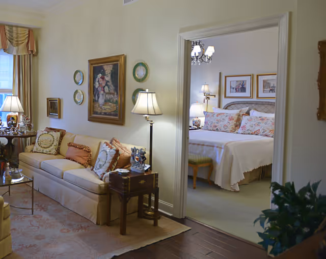 A cozy living room with a beige sofa adorned with decorative pillows, a side table with a lamp, and framed artwork on the wall. Through an open doorway, a bedroom is visible with a neatly made bed featuring floral pillows, bedside lamps, and framed pictures on the wall.