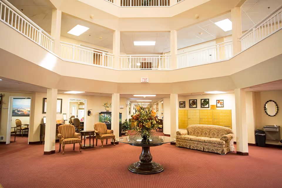 A spacious and well-lit common area in a retirement community featuring a round glass table with a floral arrangement in the center, two striped armchairs, a patterned sofa, and a wall of mailboxes. The area has a high ceiling with a balcony railing above and a red carpeted floor.