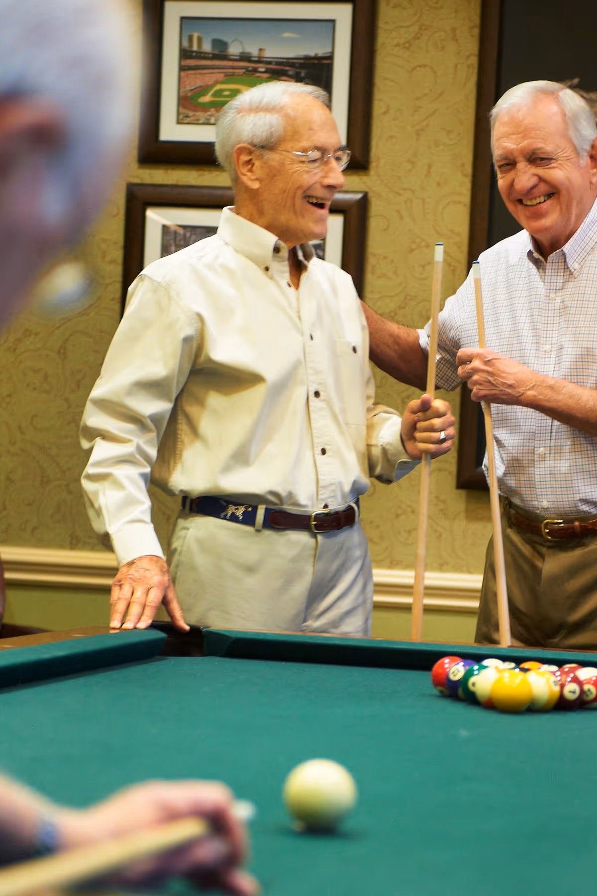 Two elderly men smiling and holding pool cues while standing next to a pool table with billiard balls arranged for a game. A third person is partially visible in the foreground, also holding a pool cue. The room has framed pictures on the wall and patterned wallpaper.