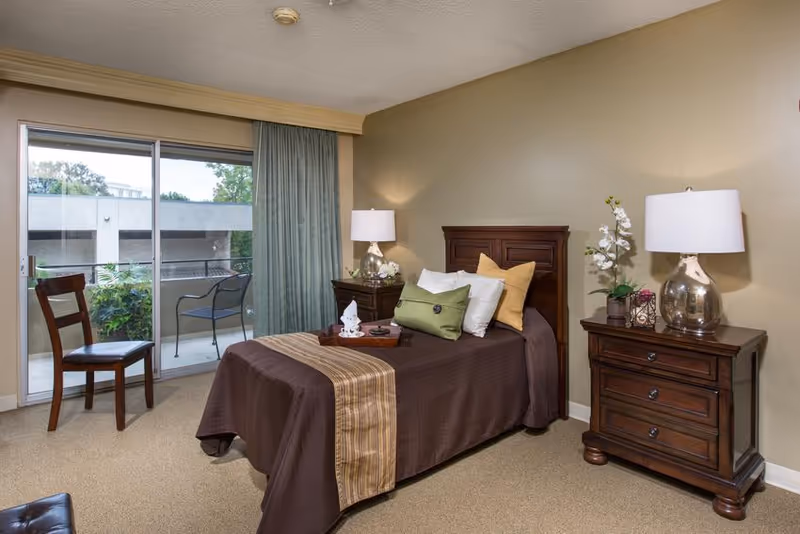 A senior living facility bedroom with a single bed covered in brown bedding and multiple pillows. There are two wooden nightstands on either side of the bed, each with a lamp and decorative items. A wooden chair is placed near a sliding glass door that leads to a balcony with outdoor seating. The walls are painted beige and the room is carpeted.