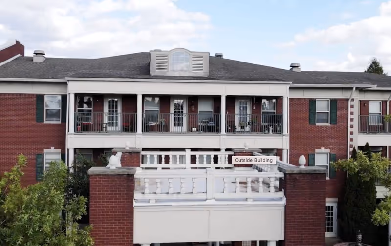 Exterior view of a two-story brick senior living community building with balconies on the upper floor, white railings, and green window shutters under a partly cloudy sky.