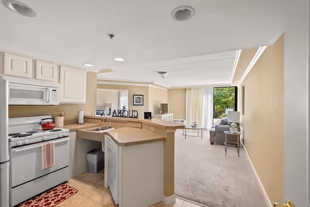 View of a kitchen area with white cabinets, a stove with a red kettle, and a dishwasher. The kitchen opens into a living room with a gray sofa, a side table with a lamp, and a large window with curtains letting in natural light.