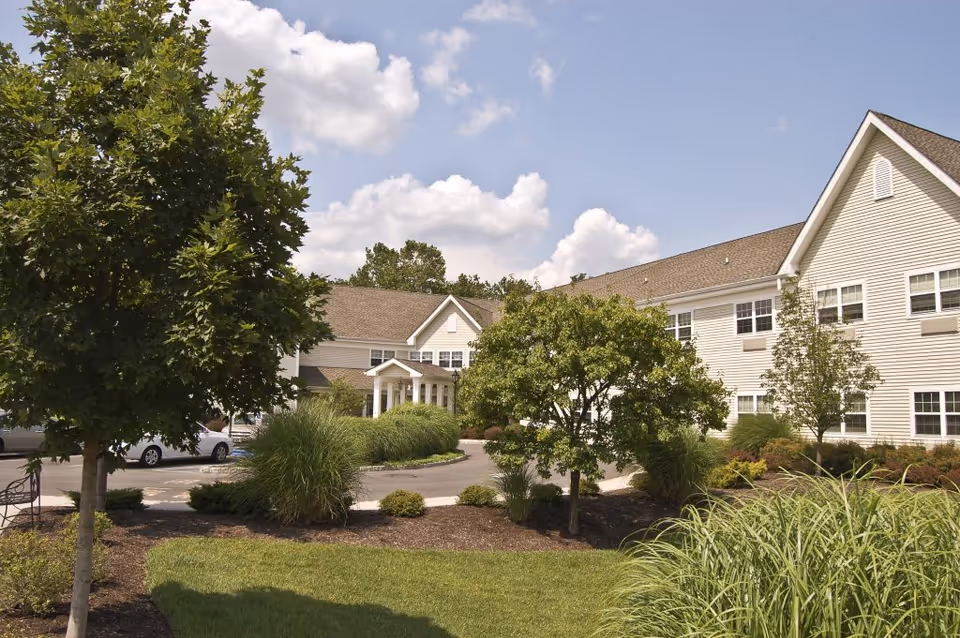 Exterior view of a two-story assisted living facility building with beige siding, surrounded by green trees, shrubs, and a well-maintained lawn under a partly cloudy sky.