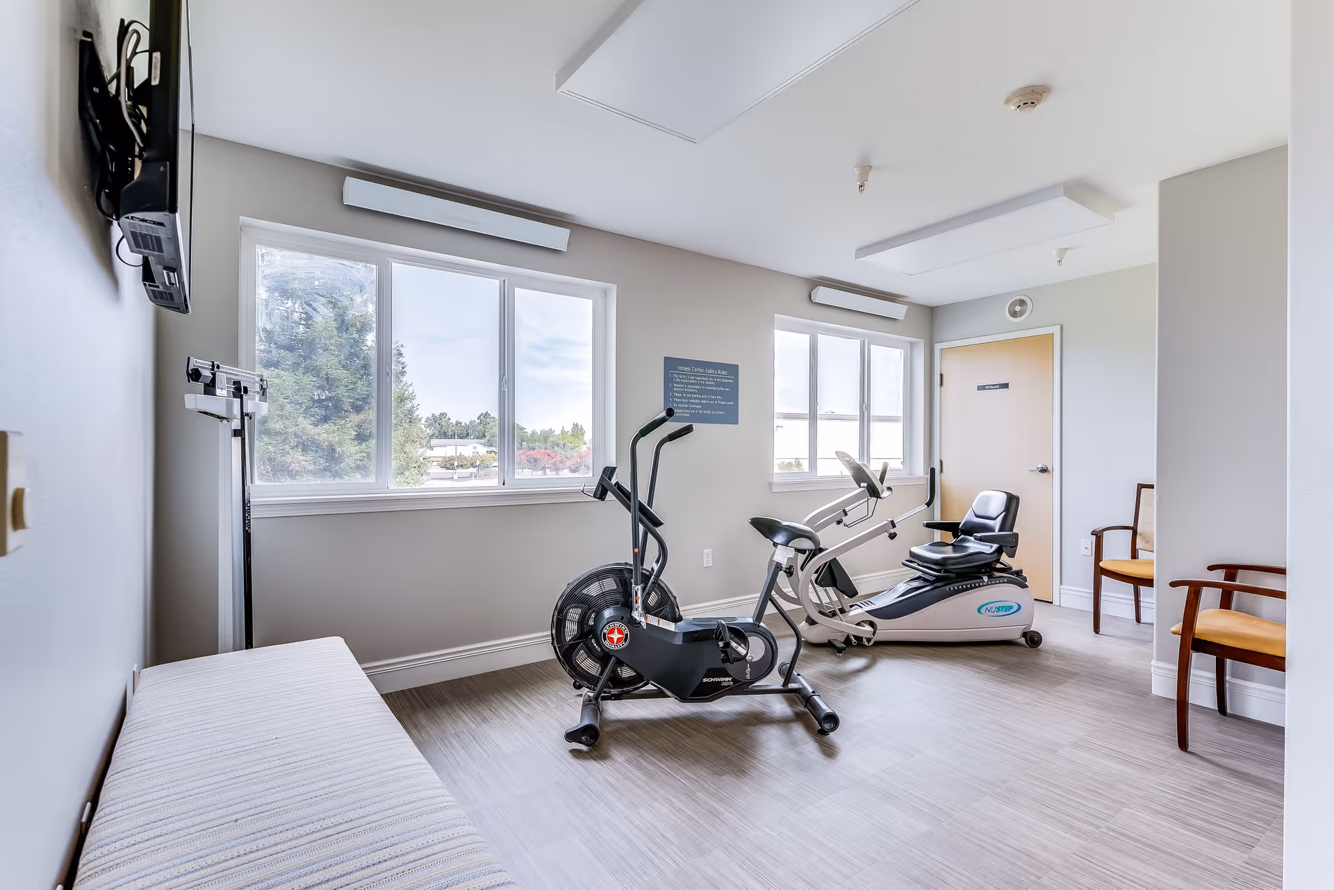 A small exercise room with two stationary bikes, a bench, two wooden chairs with yellow cushions, a wall-mounted TV, and large windows letting in natural light. The room has light-colored walls and flooring, with a door in the back corner.