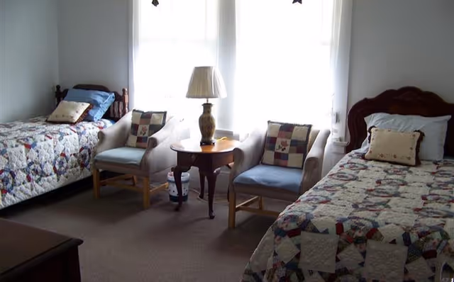 A bedroom with two twin beds, each covered with a patchwork quilt. Between the beds are two armchairs with patterned cushions and a wooden side table with a decorative lamp. The room has two large windows with sheer white curtains allowing natural light to fill the space.