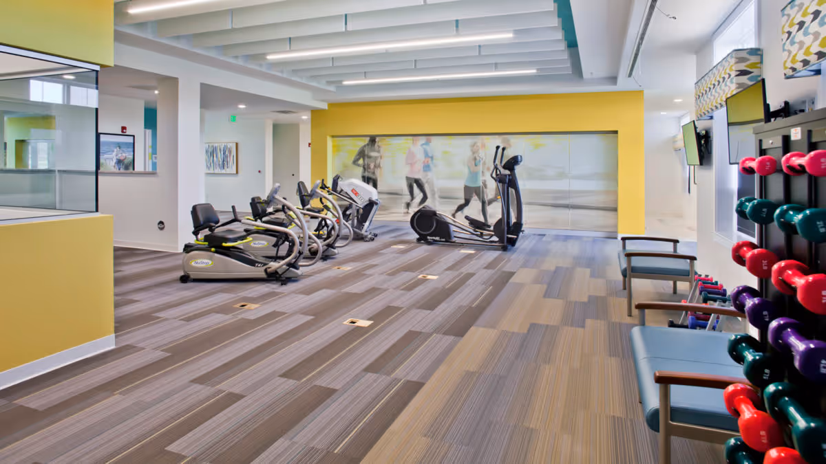 Bright senior living fitness room with cardio machines, dumbbells, and seating.