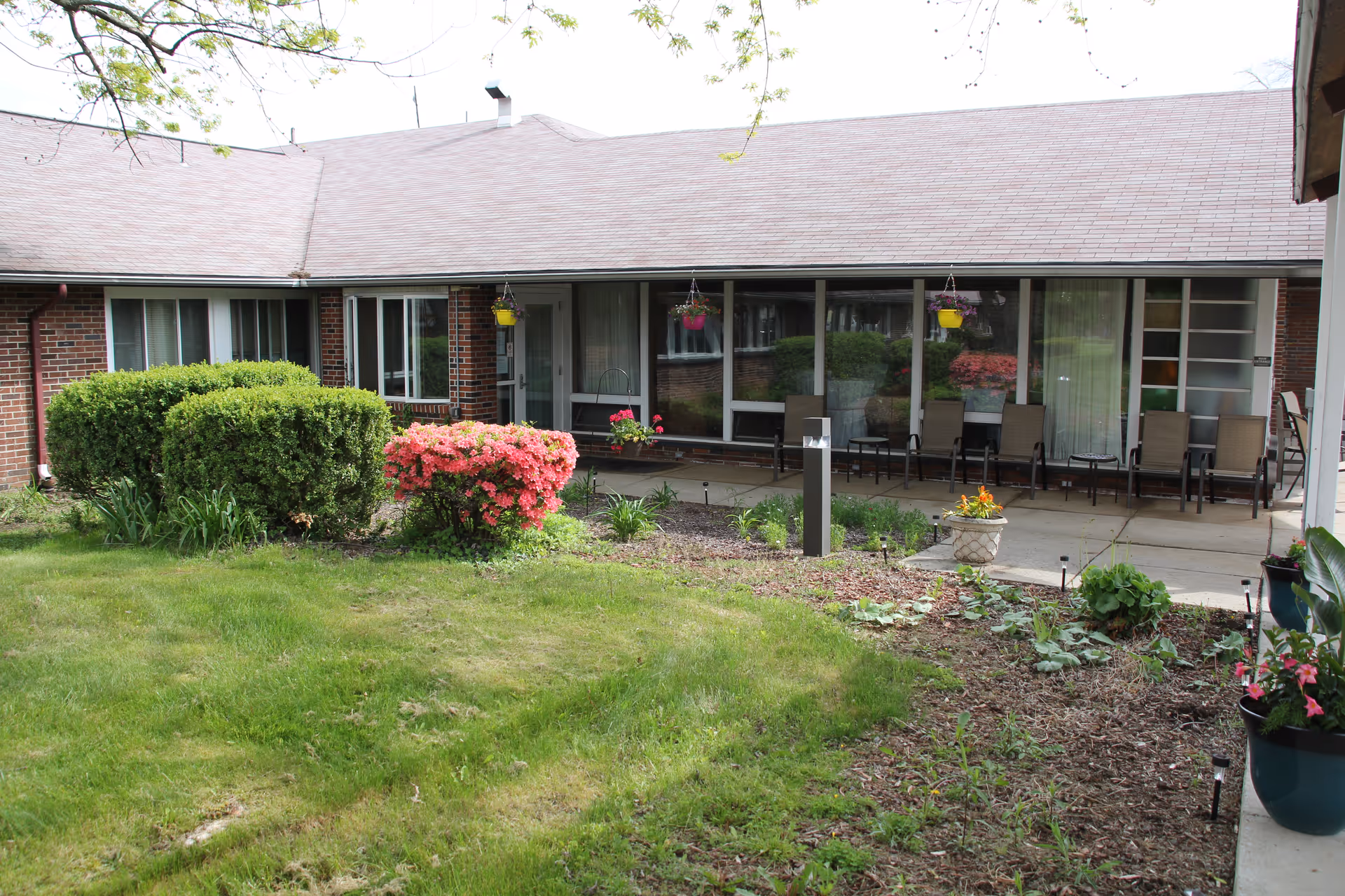 A single-story brick building with a sloped roof, large windows, and a patio area with several chairs. The foreground features a well-maintained garden with green grass, bushes, flowering plants, and hanging flower pots.
