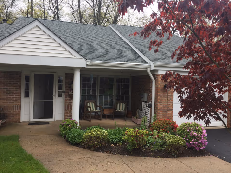 Front exterior view of a single-story brick building with a covered porch featuring two chairs with striped cushions. There is a garden with various green plants and colorful flowers in front of the porch, and a tree with red leaves on the right side. The building has a white door, windows, and a gray shingled roof.
