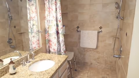 Walk-in tiled bathroom with a granite sink vanity, floral shower curtain, shower seat, grab bar, and handheld showerhead.