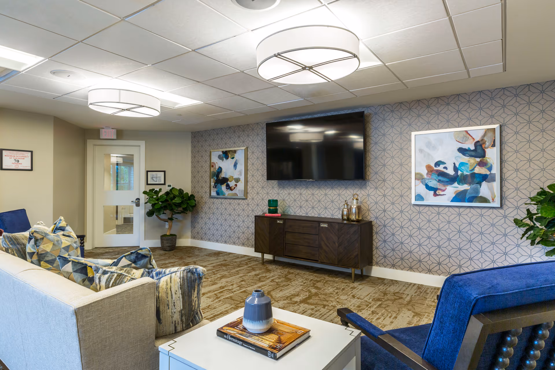 A modern living room area in Orchard Estate of Woodbury featuring a beige sofa with patterned pillows, a blue armchair, a white coffee table with a book and decorative vase, a wall-mounted flat-screen TV, two abstract paintings on a patterned wallpapered wall, and two large ceiling lights. There are also two potted plants and a door with a glass panel.