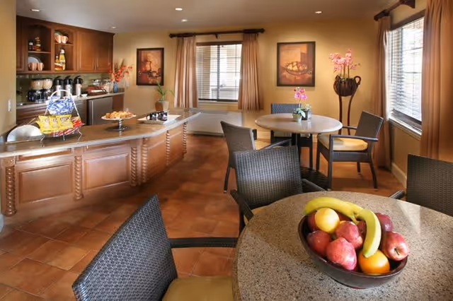 A cozy dining area with round tables and wicker chairs. One table has a bowl of fresh fruit including bananas, apples, and oranges. In the background, there is a kitchen counter with snacks and coffee dispensers. The room has warm lighting, beige curtains, and framed artwork on the walls.
