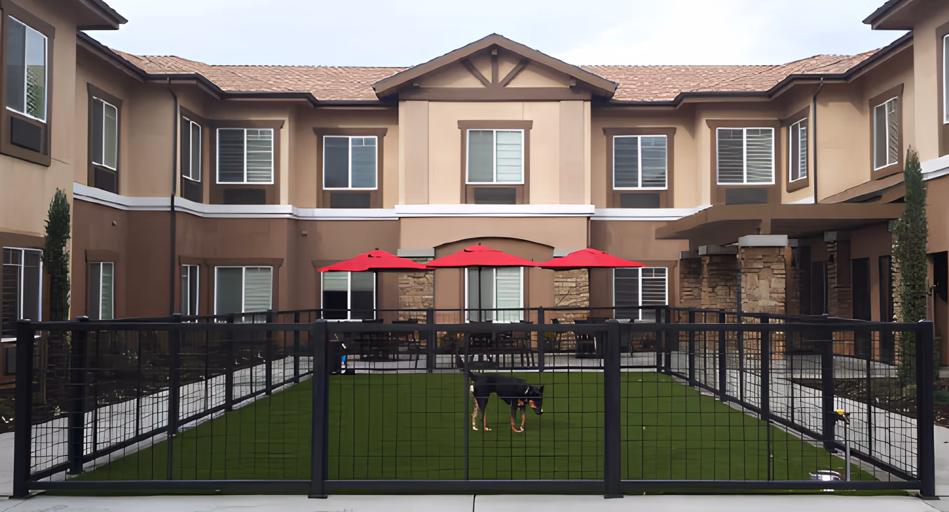 Outdoor courtyard area of Paintbrush Assisted Living with a fenced-in green artificial turf space containing a small dog. The courtyard is surrounded by a two-story building with multiple windows and three red umbrellas shading seating areas.