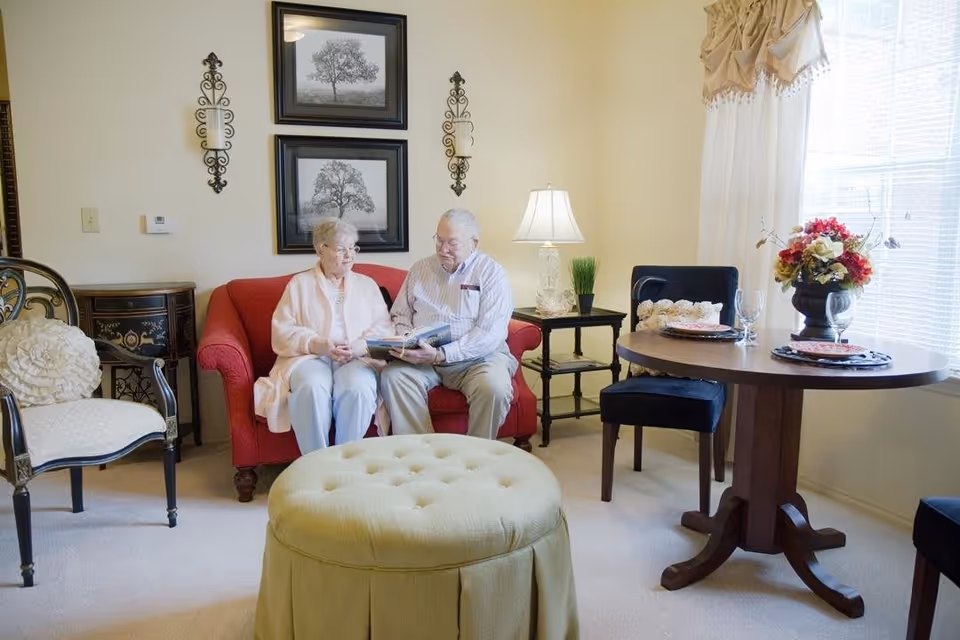 An elderly couple sitting on a red couch in a cozy living room, looking at a photo album together. The room features a round wooden dining table set with plates and glasses, a cushioned ottoman, a side table with a lamp and a small plant, and decorative wall art and candle holders. Natural light comes through a window with sheer curtains.
