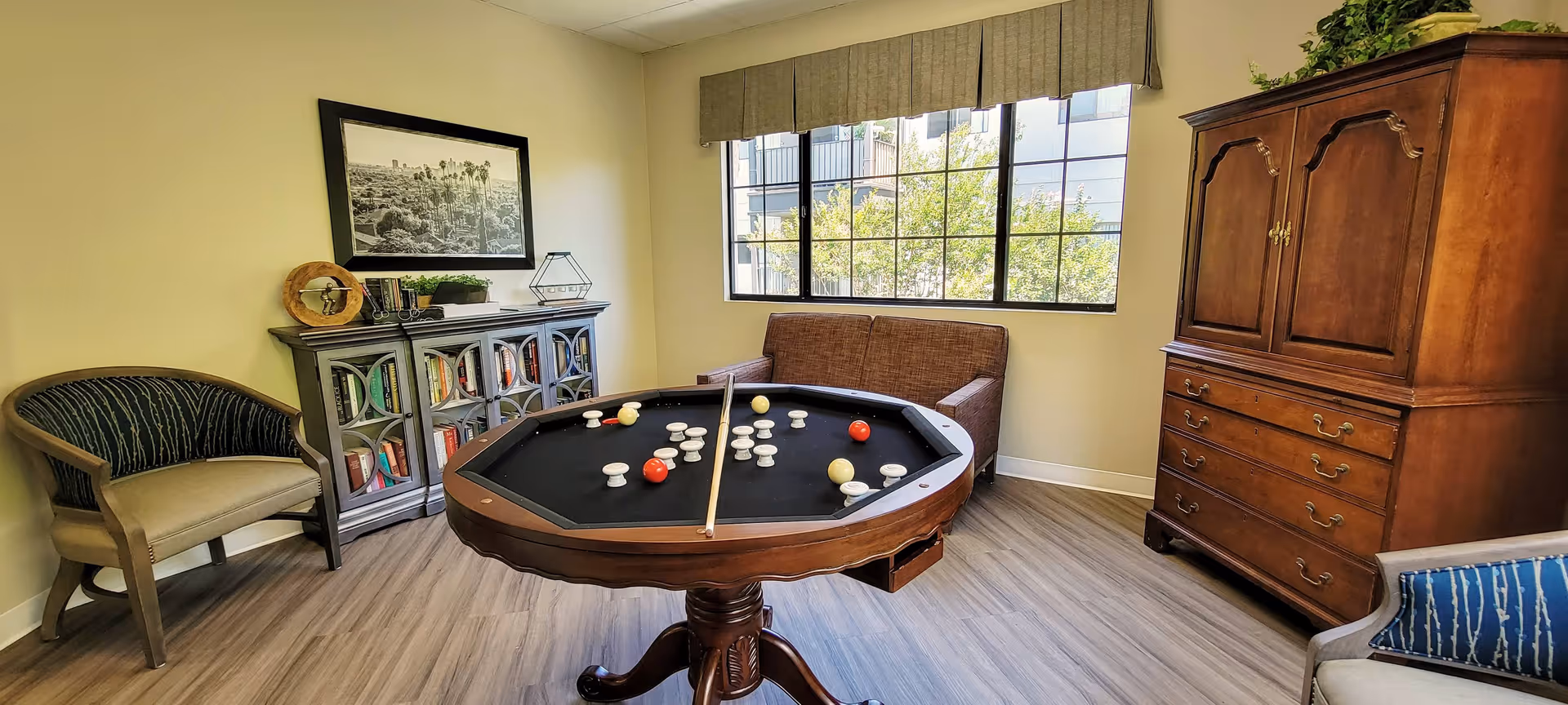 A cozy room with a round game table featuring a billiard-style game with white and red balls and a cue stick. The room has wooden flooring, a large window with a view of trees outside, a brown loveseat, two armchairs, a gray bookshelf filled with books and decorative items, and a tall wooden cabinet with drawers.