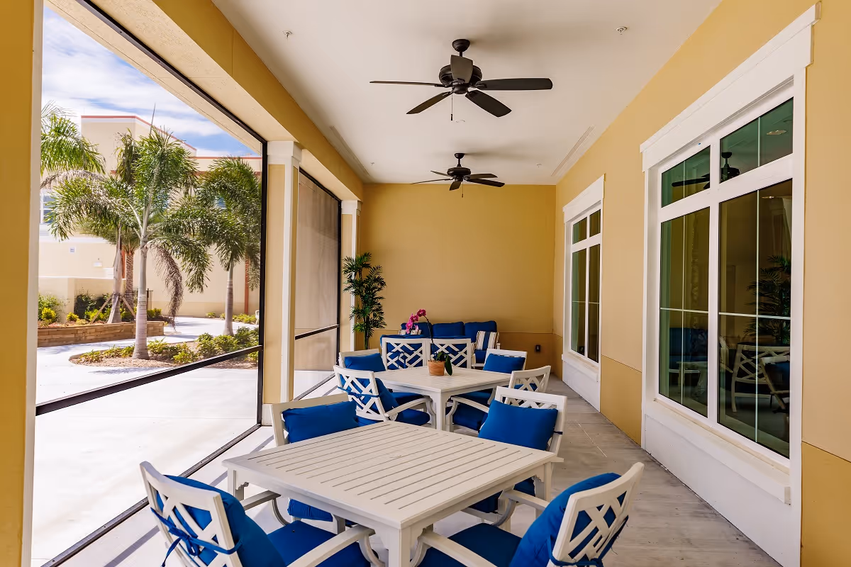A covered outdoor patio area with white tables and chairs featuring blue cushions. The patio has ceiling fans and potted plants, with a view of palm trees and a sunny sky outside.