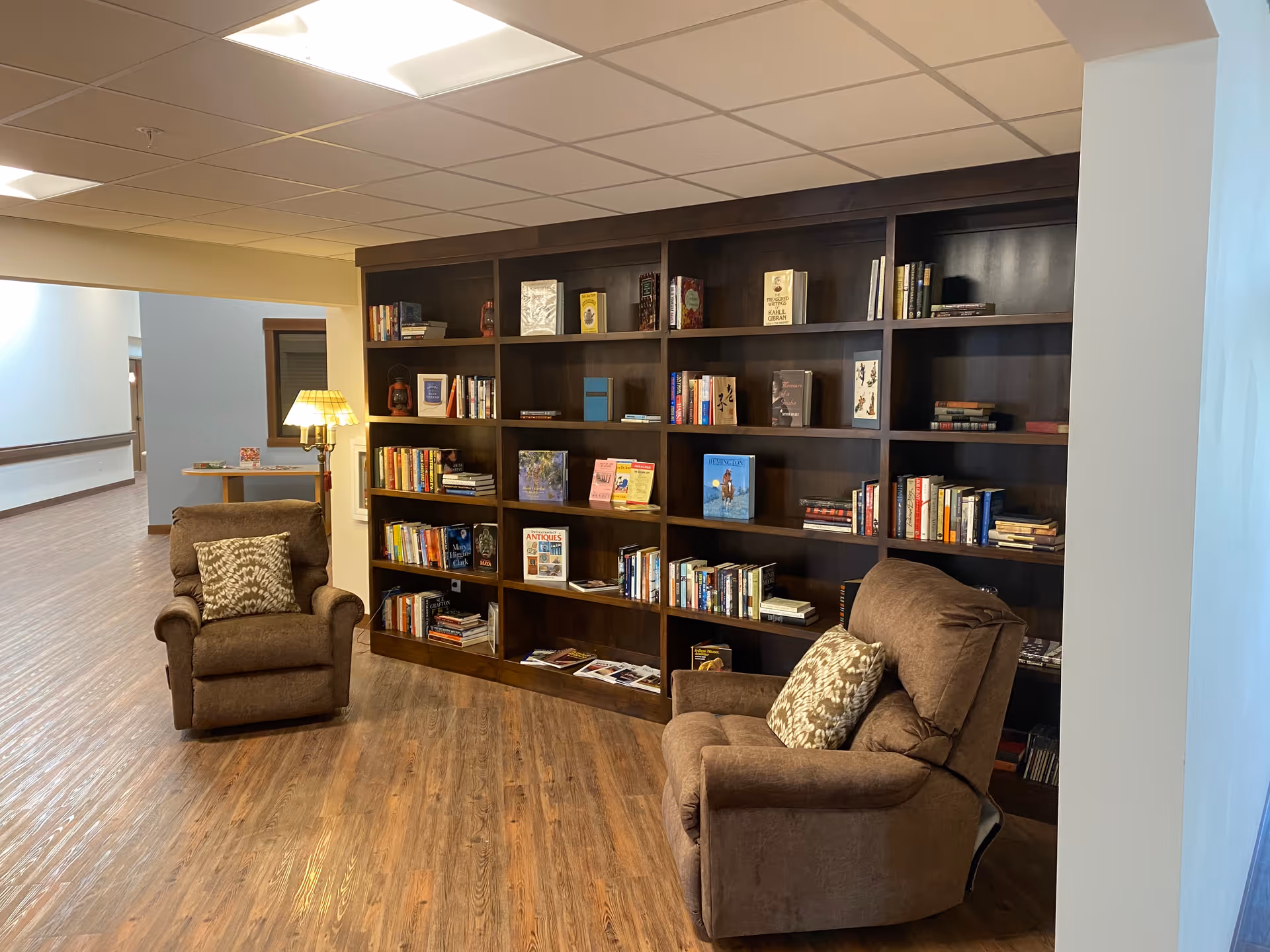 A cozy reading nook in a senior living facility featuring two brown upholstered armchairs with patterned cushions, positioned in front of a large dark wooden bookshelf filled with books and magazines. A floor lamp with a yellow lampshade provides warm lighting. The area has wood flooring and a drop ceiling with recessed lighting.