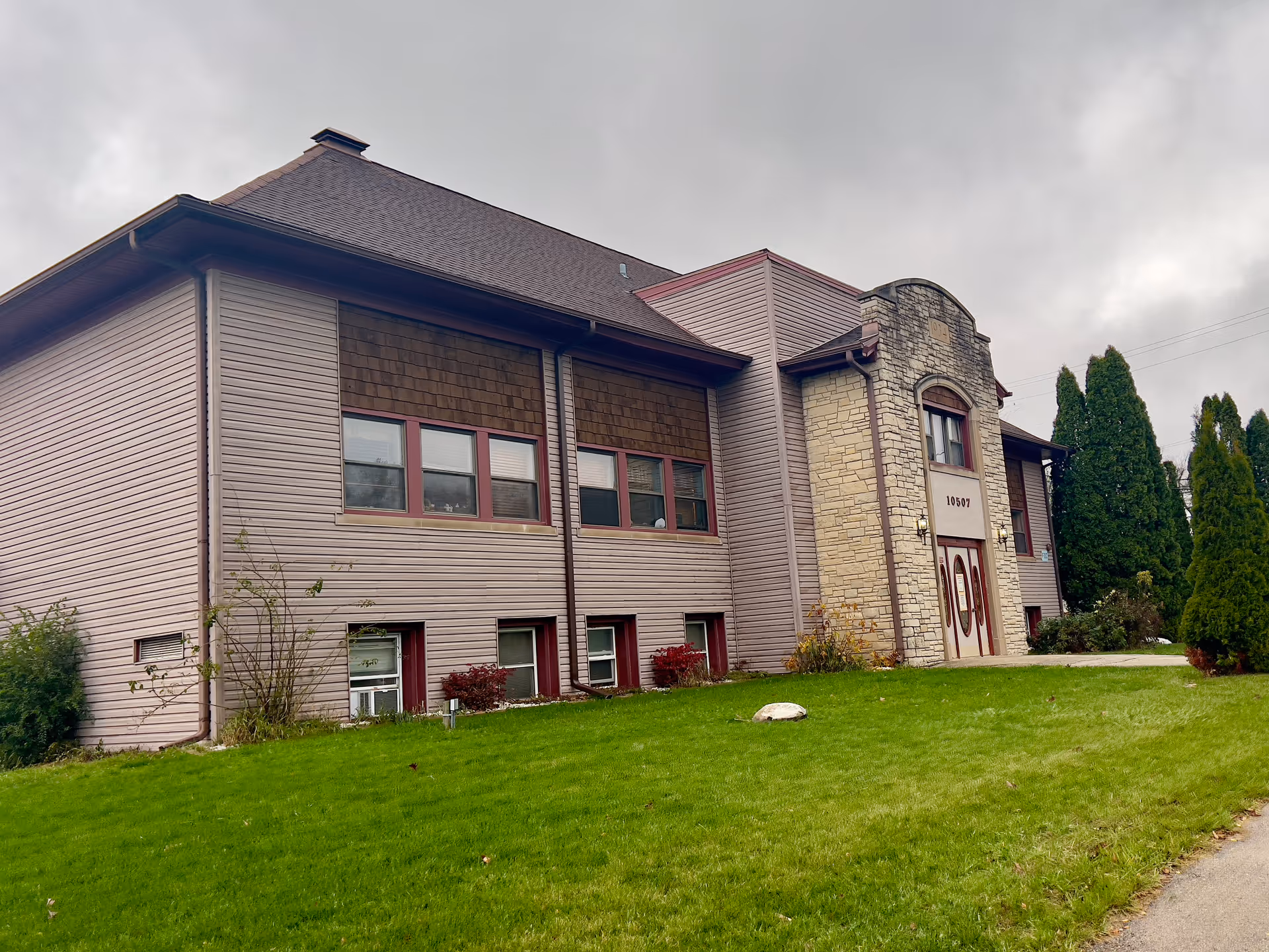 Exterior view of a two-story assisted living facility building with beige siding, brown shingles, and a stone entrance. The building has multiple windows and a green lawn in front with some bushes and trees. The sky is overcast.