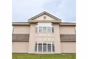 Light-colored two-story building facade with a central bay of multi-pane windows above a grassy lawn under a cloudy sky.