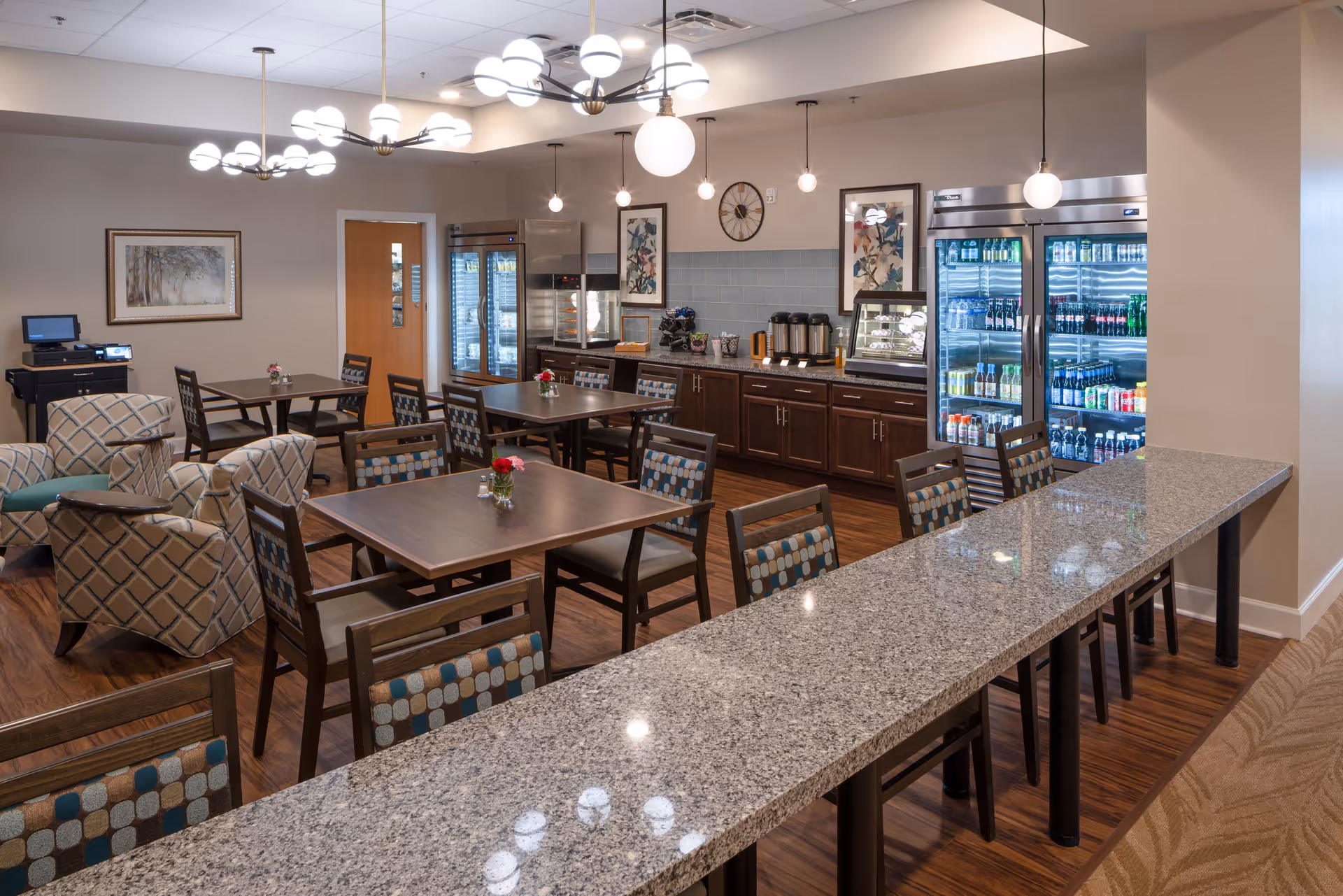 A well-lit dining area in The Culpeper facility featuring multiple wooden tables and chairs with patterned upholstery. There are comfortable armchairs in the corner, a long granite countertop with stools, and a beverage station with refrigerators stocked with drinks, coffee dispensers, and a display case. Modern pendant and chandelier lighting fixtures hang from the ceiling, and framed artwork decorates the walls.