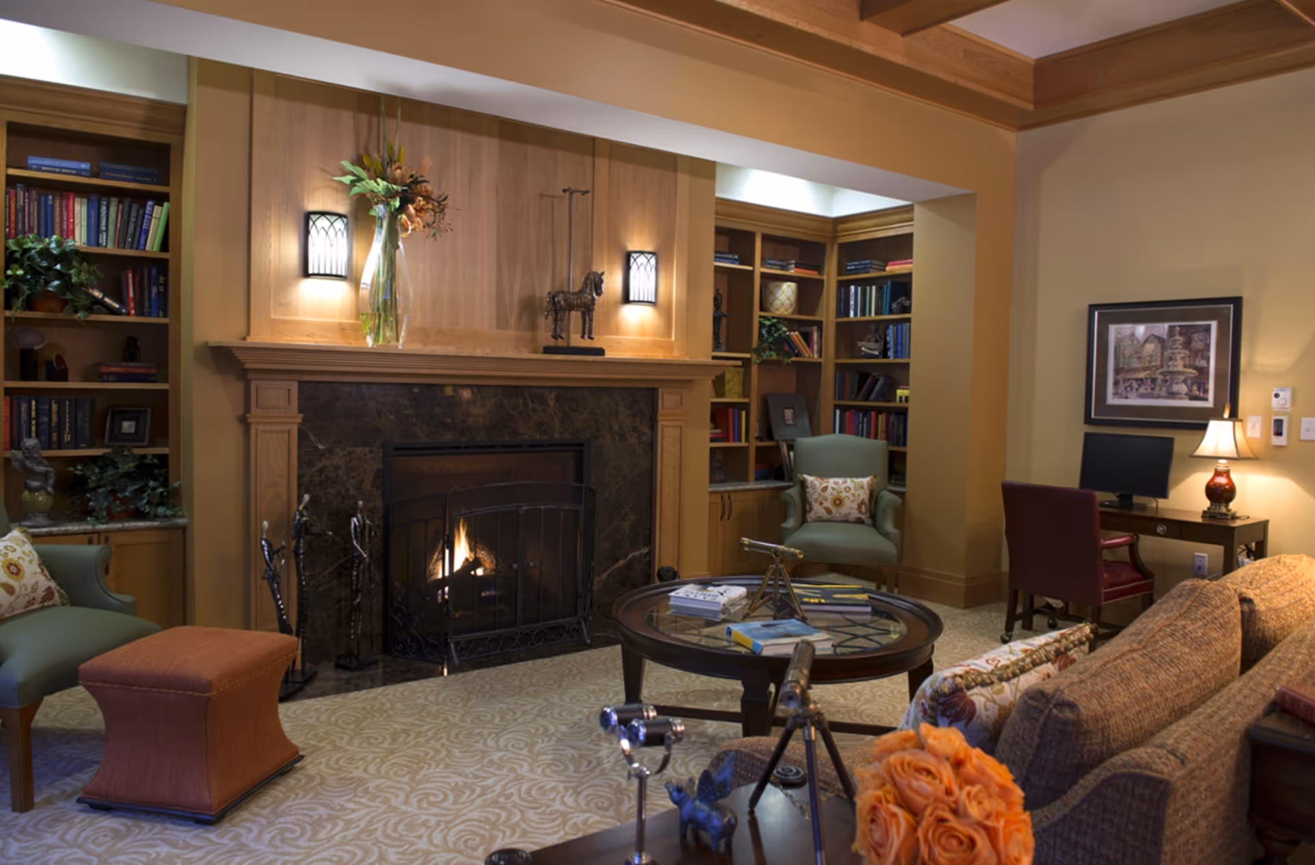 A cozy living room featuring a fireplace, comfortable seating, a round coffee table with books, and a bookshelf filled with various items. Decorative flowers and a lamp add warmth to the space.