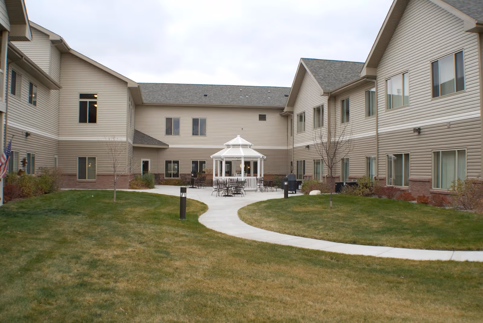 Outdoor courtyard area of Sweetwater Retirement Community featuring a white gazebo with tables and chairs around it, surrounded by a two-story beige building with multiple windows. There is a curved concrete pathway through a grassy lawn leading to the gazebo.