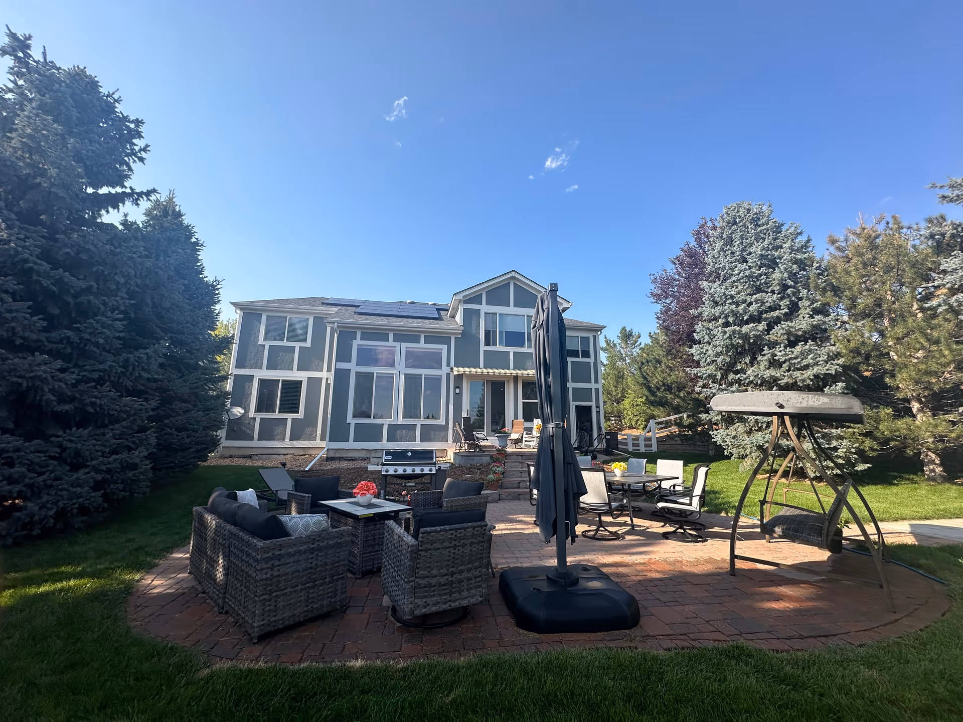 Backyard patio with wicker seating, a dining set, umbrella and swing in front of a two-story house surrounded by trees.