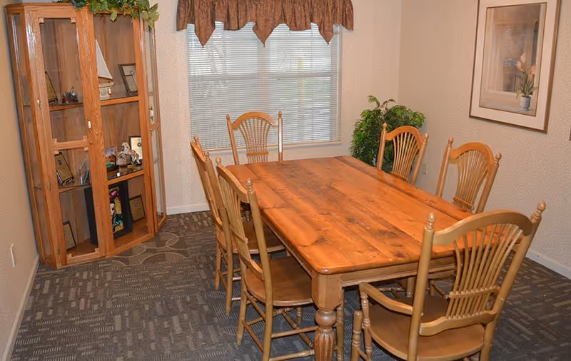 Wooden dining table with six chairs in a small dining room with a display cabinet and a window.
