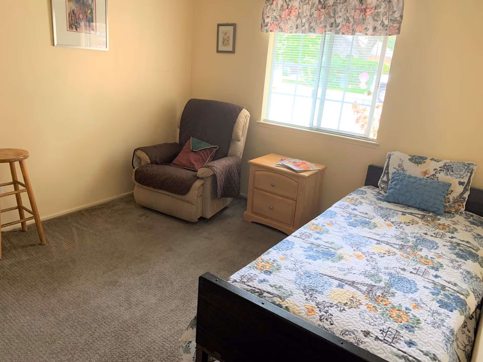 A cozy bedroom with a single bed covered in a floral quilt and a blue decorative pillow. Next to the bed is a wooden nightstand with a magazine on top. A cushioned armchair with a brown cover and two pillows sits in the corner near a window with floral curtains. A wooden stool is placed against the opposite wall, and two framed pictures hang on the walls.