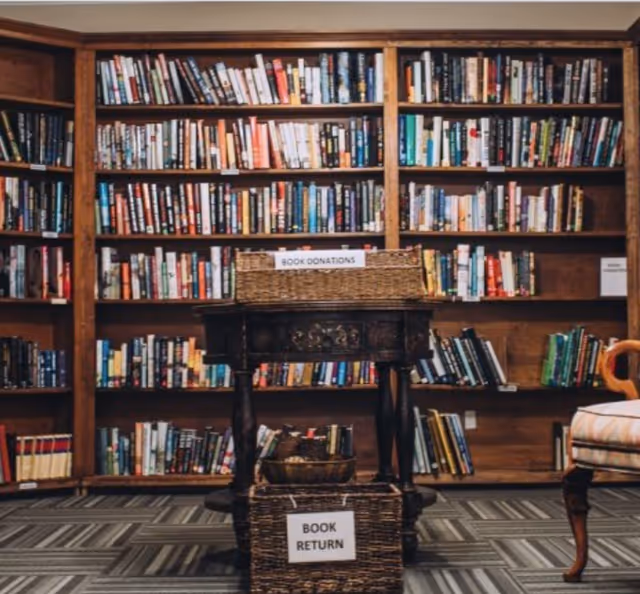 Wooden bookcases filled with books behind a table and baskets labeled 'BOOK DONATIONS' and 'BOOK RETURN' in a reading area.