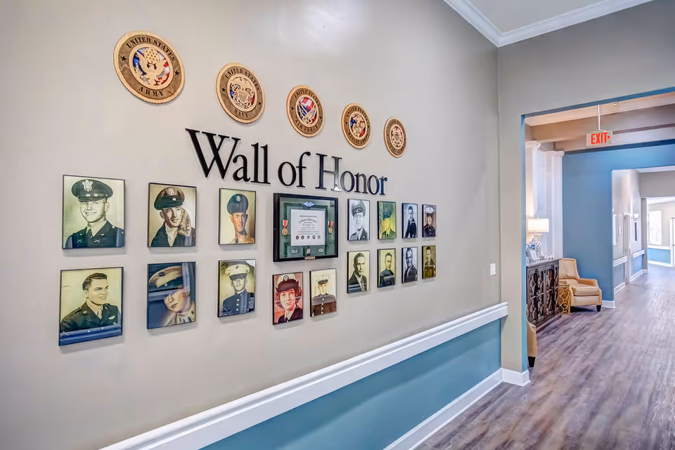 A hallway in a senior living facility with a 'Wall of Honor' display featuring framed photographs of military personnel and plaques representing different branches of the United States Armed Forces. The hallway has wood flooring, light-colored walls with a blue accent, and a seating area with a chair and lamp visible in the background.