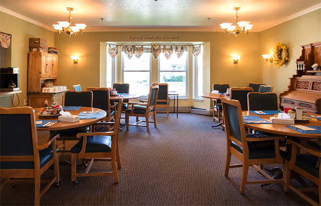 Bright assisted-living dining room with multiple round tables and wooden chairs arranged around a large bay window.