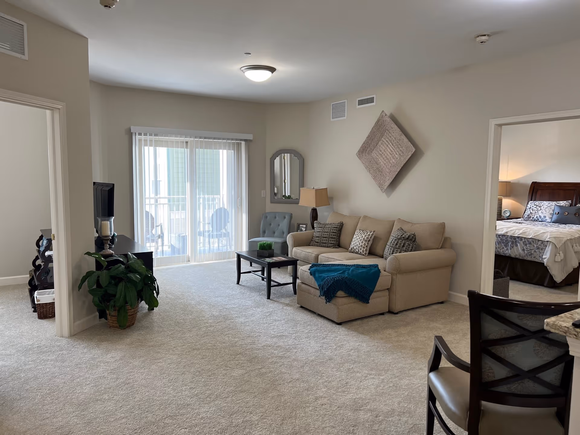 A bright living room with beige carpet and walls, featuring a beige sofa with patterned cushions and a blue throw, a black coffee table with a small plant, a gray armchair, and a wall mirror. Sliding glass doors with vertical blinds lead to a balcony with outdoor chairs. To the right, an open doorway reveals a bedroom with a bed, nightstand, and lamp. A potted plant is near the left doorway, and a chair with a dark wooden frame is partially visible in the foreground.