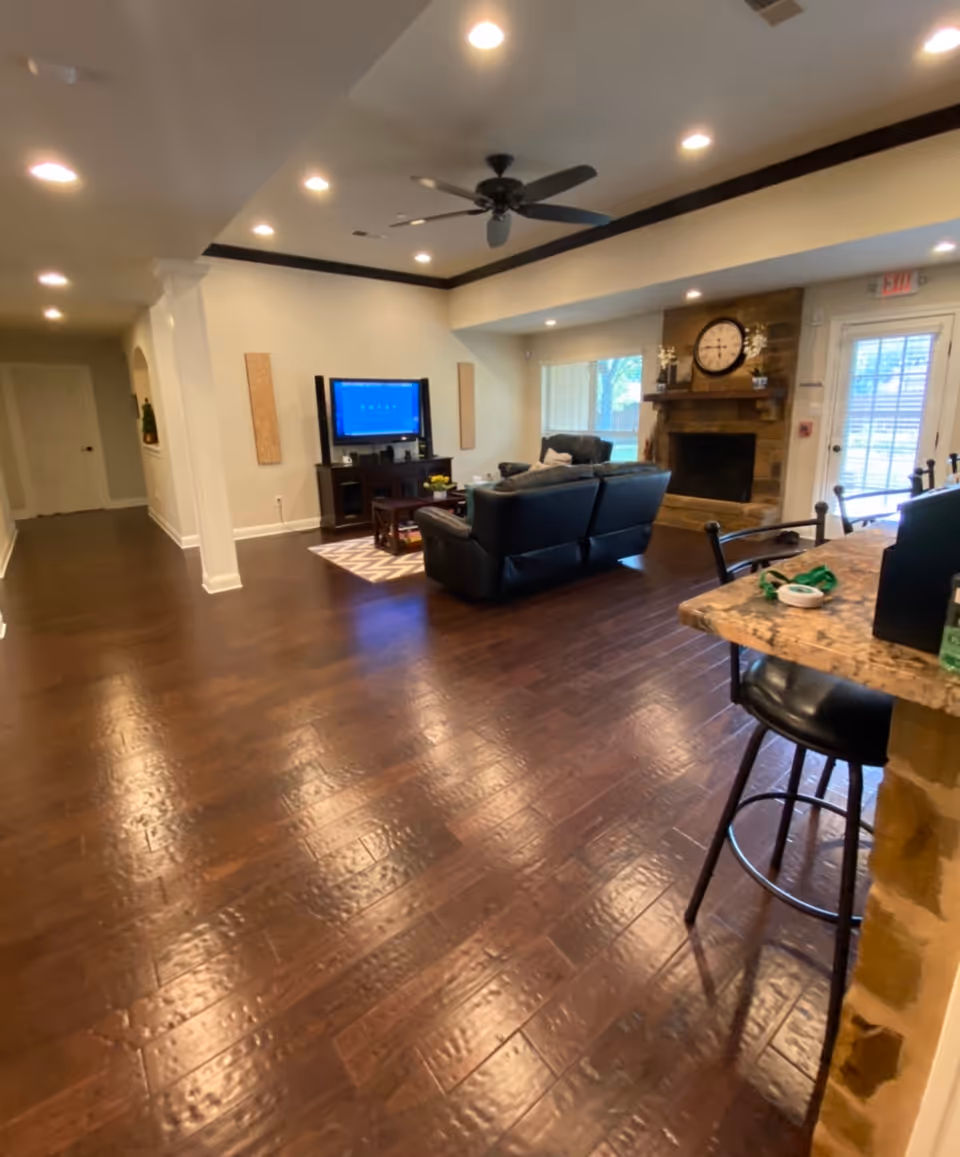 Open living room with dark hardwood floors, leather sofas facing a TV and a stone fireplace with a clock, and a bar counter with stools in the foreground.