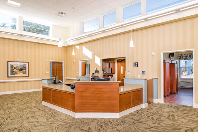 A spacious reception area with a large wooden desk in the center, beige patterned wallpaper, carpeted floor with a leafy design, and several pendant lights hanging from a high ceiling with skylights. There are doorways leading to other rooms, and a framed painting on the wall.