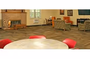Carpeted communal lounge with a brick fireplace, upholstered chairs, and a round table with red chairs in the foreground.