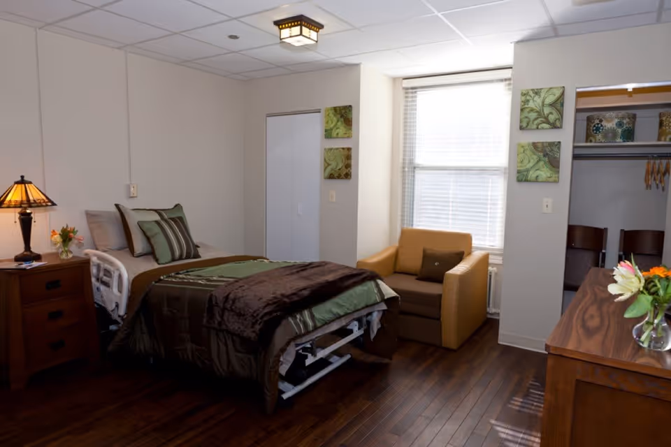 A furnished bedroom with a made bed, nightstand and lamp, armchair, dresser and a window.