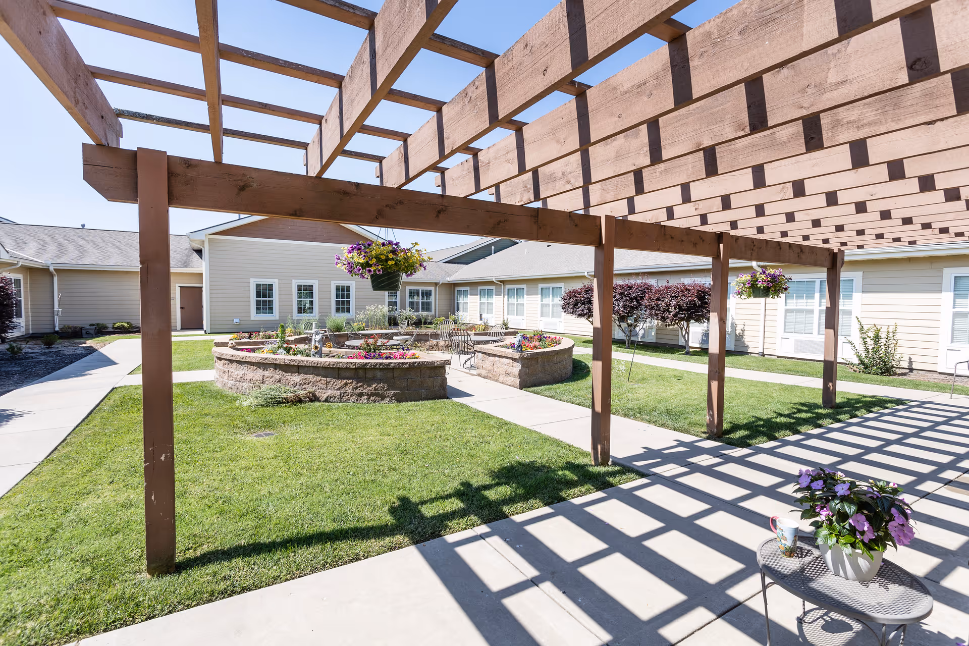 Outdoor courtyard area at Oak Pointe of Neosho featuring a wooden pergola casting shadows on a concrete walkway. There are hanging flower baskets and a small round table with a potted plant and a cup. In the background, there are raised circular flower beds with colorful flowers and a building with beige siding and white-framed windows.