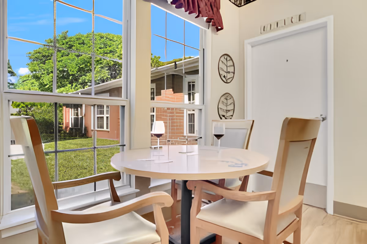 A small round dining table with four wooden chairs around it, two glasses of red wine on the table, large windows showing green trees and neighboring buildings outside, and decorative wall art with leaf designs on a light-colored wall.
